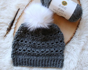 May include: A gray crocheted beanie with a white faux fur pom pom. The beanie is made with a lacy stitch pattern. The yarn is labeled "Jeans Jean Vaqueros".