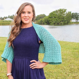 Könnte beinhalten: Eine Frau trägt ein blaues Kleid mit einer türkisfarbenen Häkeljacke. Die Jacke hat ein Spitzenmuster und ist vorne offen. Die Frau steht auf einer grünen Wiese mit einem See im Hintergrund.