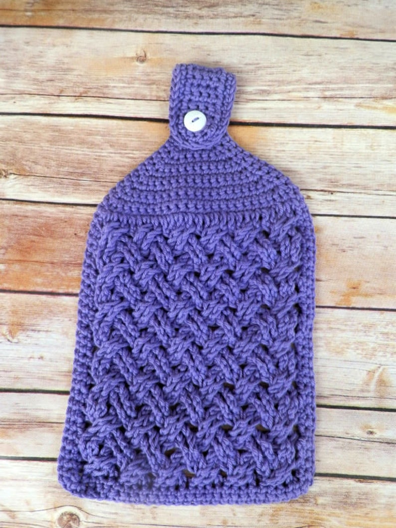 Puede incluir: Un pa&ntilde;o de cocina de ganchillo morado hecho a mano con cierre de bot&oacute;n. La toalla presenta un patr&oacute;n tejido texturizado y un lazo para colgar. El bot&oacute;n es blanco y redondo. La toalla se muestra sobre una superficie de madera marr&oacute;n claro.