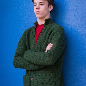 May include: A young person wearing a green knitted cardigan with a red t-shirt underneath. The cardigan has a button closure and two pockets. The person is standing against a blue wall.