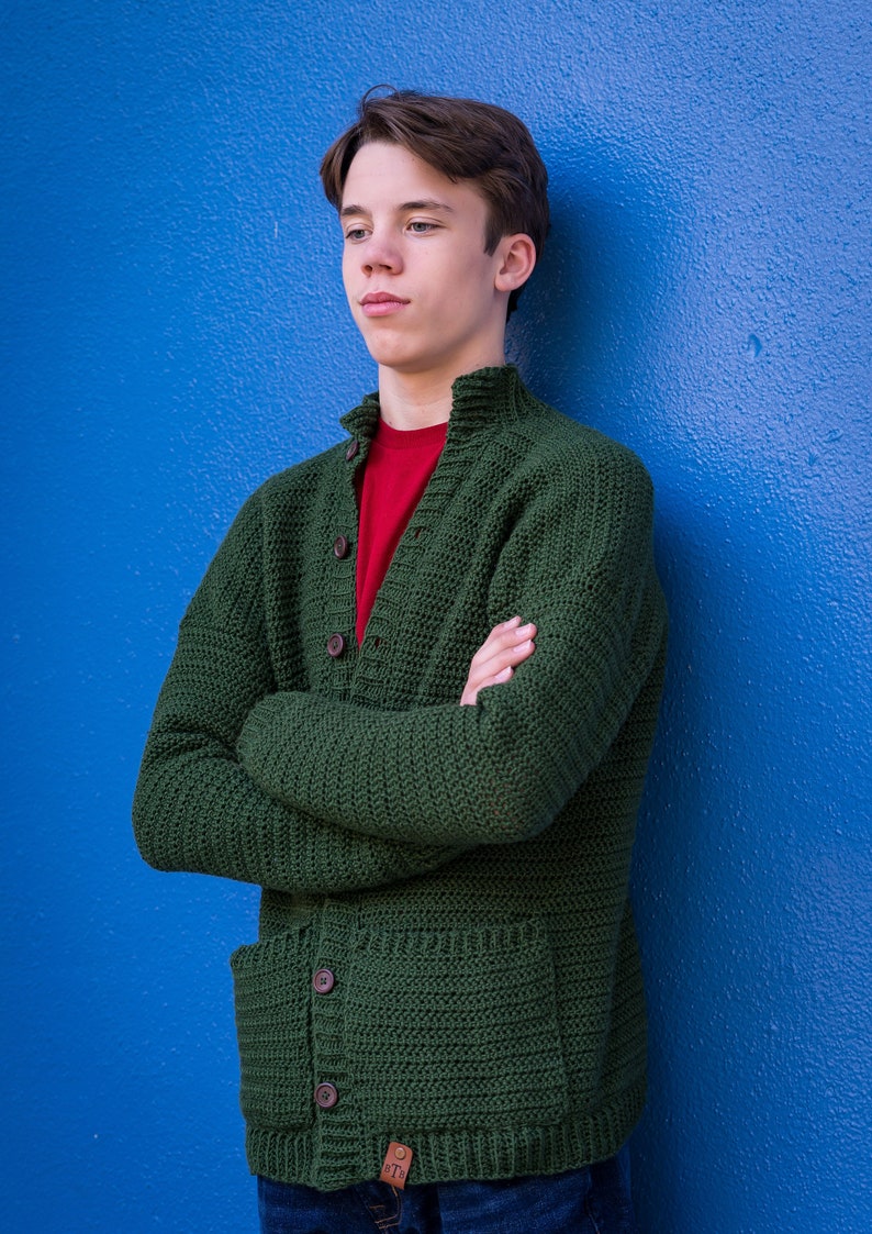 May include: A young person wearing a green knitted cardigan with a red t-shirt underneath. The cardigan has a button closure and two pockets. The person is standing against a blue wall.