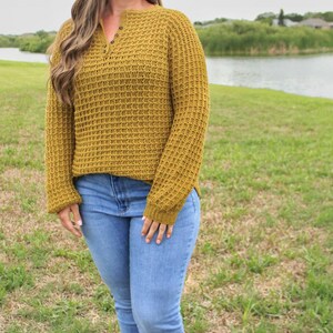 May include: A mustard yellow waffle knit sweater with a single button closure at the neckline. The sweater is worn with light blue jeans.
