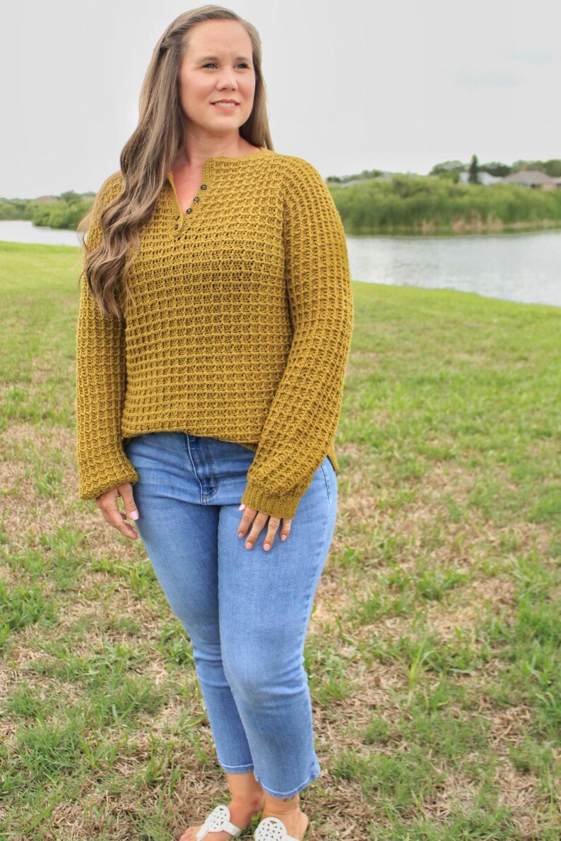 May include: A mustard yellow waffle knit sweater with a single button closure at the neckline. The sweater is worn with light blue jeans.