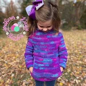 May include: A young person wearing a purple, blue, and pink striped crocheted sweater with a white and purple bow in their hair. The sweater has a large pocket on the front.