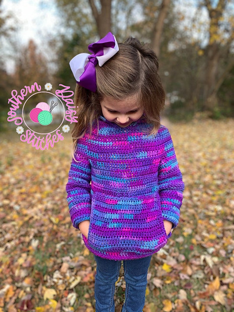 May include: A young person wearing a purple, blue, and pink striped crocheted sweater with a white and purple bow in their hair. The sweater has a large pocket on the front.