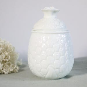 Peut inclure: Un pot en céramique blanche avec couvercle, présentant un motif texturé en relief rappelant les écailles d'un ananas sur toute sa surface. Le pot possède un corps arrondi et un petit bouton décoratif sur le couvercle, avec une finition émaillée.