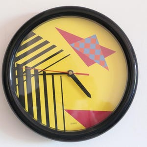 May include: A black framed clock with a yellow face and geometric designs in red, blue, and black. The clock hands are black.