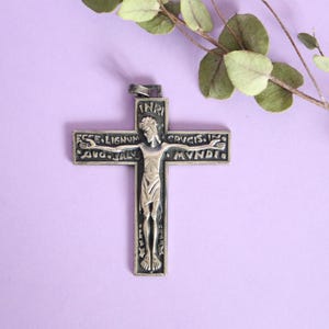 May include: Silver-toned crucifix pendant with a detailed depiction of Jesus Christ. The cross features engraved Latin text and is displayed against a soft purple background, with a sprig of greenery in the upper right corner.
