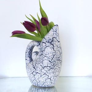 May include: A white and navy blue crackle-glazed ceramic vase with a handle, holding several purple tulips with green leaves. The vase has a unique, organic shape and sits on a reflective surface.