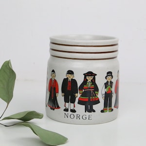 Peut inclure: Un pot en céramique avec une finition mouchetée beige clair. Le pot est décoré d'illustrations de personnes en vêtements traditionnels norvégiens. Le mot "NORGE" est imprimé en noir sur le devant du pot.