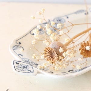 May include: A white ceramic dish with a blue floral pattern holds dried flowers and seed pods. The dish has a scalloped edge and a handle. The arrangement is set against a light background.