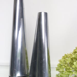 May include: Two polished silver vases with a conical shape. The vases are displayed on a reflective surface, with a light green floral arrangement in the background. The vases have a sleek, modern design.