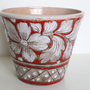 May include: A red and white ceramic planter with a floral design. The planter has a wide mouth and a narrow base.