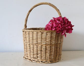 Könnte beinhalten: Ein hellbrauner Weidenkorb mit gebogenem Griff, gefüllt mit leuchtend rosa Blüten. Der Korb ist rund und gewebt. Der Griff besteht aus gedrehtem Weidengeflecht. Die Blumen sorgen für einen Farbtupfer.