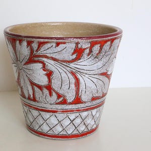 Puede incluir: Una maceta de cerámica con interior beige y exterior blanco con un diseño floral rojo. La maceta tiene un borde y una base rojos, con un patrón de diamantes cerca de la parte inferior. La maceta mide aproximadamente 15 cm de altura.