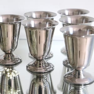 Könnte beinhalten: Fünf silberne Metallbecher mit geriffeltem Design und runder Basis. Die Becher sind in einer Reihe auf einer reflektierenden Oberfläche angeordnet.