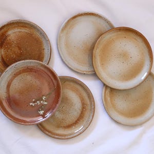 以下が含まれることがあります： 斑点のある釉薬で覆われた、茶色と白の様々な色合いのセラミックプレート6枚セット。プレートは丸く、わずかに盛り上がった縁があります。
