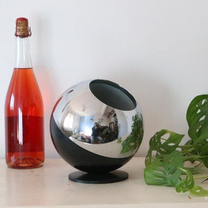 Può includere: Una bottiglia di vetro trasparente riempita con un liquido rosso, accanto a un oggetto decorativo sferico cromato e nero. Una pianta verde con motivi fogliari unici è sullo sfondo.