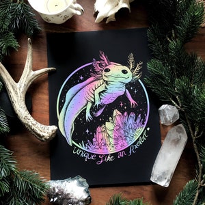 Puede incluir: Una impresión en blanco y negro de un ajolote rodeado de cristales. El ajolote es iridiscente y tiene un esquema de color rosa y azul. El texto "Unique Like an Axolotl" está escrito debajo del ajolote.