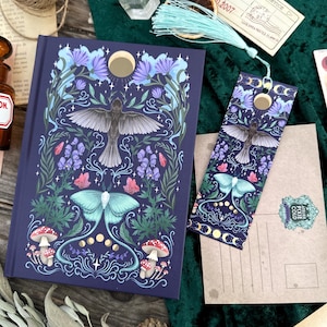 May include: A dark blue journal and bookmark set with a nature-inspired design. The cover features a central bird image with floral and mushroom illustrations in shades of blue, purple, and red. A gold moon accentuates the design.