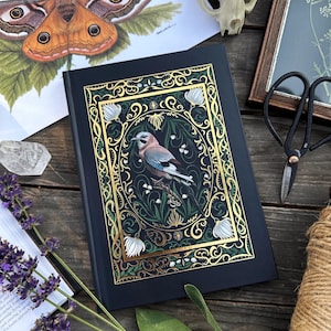 Könnte beinhalten: Ein schwarzes Hardcover-Journal mit einem goldenen und grünen floralen Design, das einen Blauhäher zeigt. Das Journal liegt auf einer Holzoberfläche mit anderen Dekorationsartikeln.