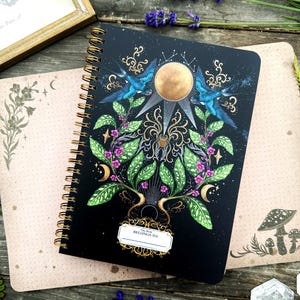 May include: A black spiral bound journal with a gold and black design featuring a sun, birds, and a plant with flowers. The text "This Book Belongs To" is on the front cover.