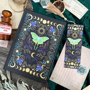 Puede incluir: Un diario negro y un marcapáginas a juego presentan un diseño celestial con una polilla verde, lunas doradas y detalles morados. El diario está cerrado y se encuentra junto a una postal y otros elementos decorativos.