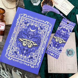 May include: A purple journal and bookmark set with a gold moth and celestial design. The journal has a gold border and intricate details. A purple tassel hangs from the bookmark. The items are on a green velvet surface, with other decorative items.