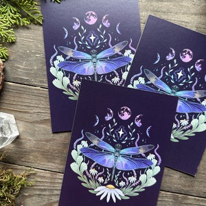May include: Three art prints featuring a detailed illustration of a dragonfly with iridescent blue wings. The design includes phases of the moon, stars, and floral elements, set against a dark purple background. The prints are displayed on a rustic wooden surface.