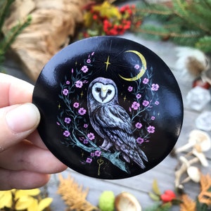 Può includere: Un gufo bianco e nero con una luna crescente gialla e fiori rosa su uno sfondo nero. Il gufo è appollaiato su un ramo con un buco della serratura sullo sfondo.