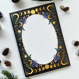 May include: A black and gold notepad with a moon and star design. The notepad has a dotted interior and a gold oval frame.