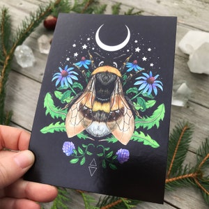 May include: A black card featuring a detailed illustration of a bumblebee surrounded by flowers and a crescent moon. The bumblebee is yellow and black with a furry body. The flowers are blue and purple with green leaves. The background is black with white stars.