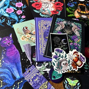 Op de afbeelding: Een verzameling briefpapierartikelen, waaronder een paars hardcover boek, een bladwijzer met een lavendelkleurig kwastje, kunstprints met een vos, walvis en kat, plus stickers van een egel, een mot en een kaars met bloemmotieven.