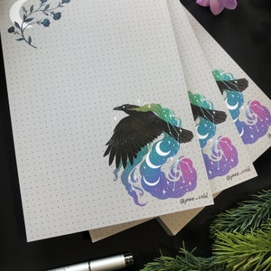 Könnte beinhalten: Drei graue und weiße Notizbücher mit Punkten und einem Design mit einem schwarzen Vogel und einem Halbmond. Das Design zeigt einen schwarzen Vogel mit einem grün-blauen Nebel dahinter. Die Notizbücher haben den Text "@pixie_cold" in der unteren rechten Ecke.