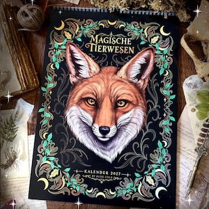 Könnte beinhalten: Ein Wandkalender für 2027 mit schwarzem Hintergrund und einer zentralen Illustration eines Fuchskopfes. Der Text lautet Magische Tierwesen und Kalender 2027 by Pixie Cold. Der Fuchs ist von einem dekorativen Rand aus grünen Blättern und gelben Monden umgeben.