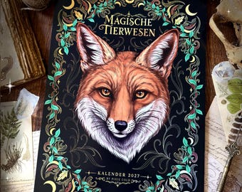 May include: A 2027 wall calendar with a black background featuring a central illustration of a fox head. The text reads Magische Tierwesen and Kalender 2027 by Pixie Cold. The fox is surrounded by a decorative border of green leaves and yellow moons.