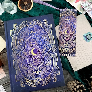 Könnte beinhalten: Ein dunkelblaues Notizbuch und ein Lesezeichen mit goldenen Himmelsmustern. Das Notizbuch zeigt einen Mond, eine Sonne und florale Muster. Das Lesezeichen hat ein passendes Design und eine lila Quaste. Weitere Artikel sind Postkarten, Kristalle und eine getrocknete Orangenscheibe.