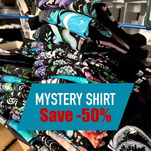 Puede incluir: Una pila de camisetas negras dobladas con varios diseños gráficos coloridos, incluyendo una luna creciente, una serpiente y patrones florales. Una pancarta turquesa muestra "MYSTERY SHIRT Save -50%" en texto blanco y rojo, anunciando un descuento.
