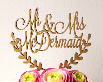 May include: A gold wooden cake topper with the text "Mr. & Mrs. McDermaid" in a cursive font. The topper is decorated with a leafy wreath and sits on top of a white cake decorated with pink and yellow flowers.