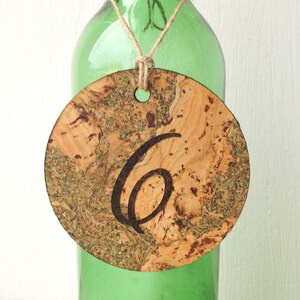 May include: A round cork place card with the number "6" engraved on it. The place card is attached to a green glass bottle with twine.