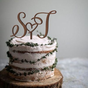 Könnte beinhalten: Ein hölzerner Kuchentopper mit den Buchstaben "S" und "T", die mit einem Herzen in der Mitte ineinander verschlungen sind. Der Topper befindet sich auf einem weißen Kuchen, der mit Grünzeug verziert ist.