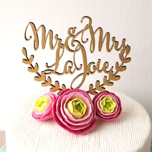 May include: A white wedding cake with a gold wooden topper that says "Mr. & Mrs. LaJoie" and three pink and yellow flowers.