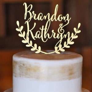 May include: Gold wedding cake topper with the names "Brandon & Kathryn" in cursive script, surrounded by a leafy wreath.