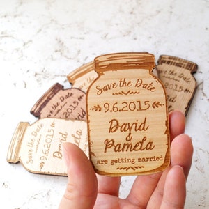 May include: Four wooden save the date magnets shaped like mason jars. The magnets are engraved with the text "Save the Date" and the date "9.6.2015". The names "David & Pamela" are engraved below the date, along with the text "are getting married".