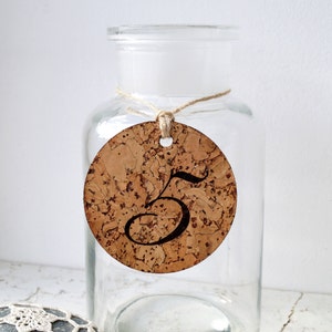 May include: A cork circle with the number 5 printed on it, hanging from a glass jar with twine.