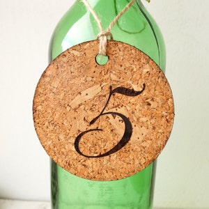 May include: Cork circle with the number 5 engraved on it. The circle is attached to a green glass bottle with twine.