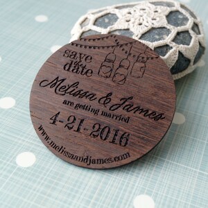May include: A round wooden save the date magnet with the text "Save the date" and "Melissa & James are getting married 4-21-2016" and the website address "www.melissaandjames.com". The magnet features a design of mason jars with hearts and string lights.