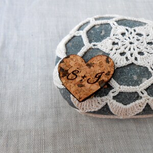 May include: A heart-shaped cork with the initials "S+J" engraved on it, placed on a gray stone with a white crocheted doily.