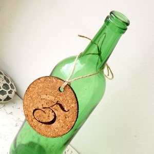 May include: A green glass bottle with a cork circle attached to it with twine. The cork circle has the number "3" written on it in cursive.