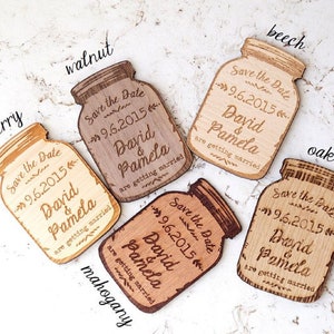 May include: Six wooden save the date magnets shaped like mason jars with the text "Save the Date" and the date "9.6.2015" engraved on them. The names "David & Pamela" and the phrase "are getting married" are also engraved on each magnet. The magnets are in different wood tones: cherry, walnut, beech, oak, and mahogany.